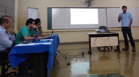 Mestrando Fabrício Vieira Campos, em pé, de camisa azul e calça preta, cabelo preto e curto, barba cerrada. Três professores avaliadores sentados: Aladir, Rodrigo Marinho e Paulo Henrique